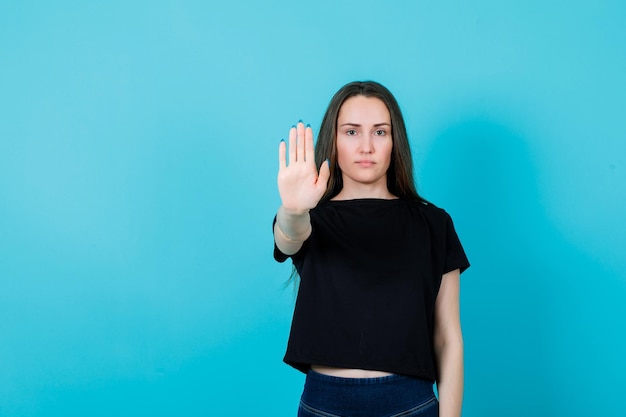 Seriously girl is showing stop gesture by extending hand to camera on blue background