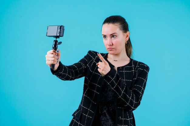 Seriously girl is taking selfie by extending forefinger to camera on blue background