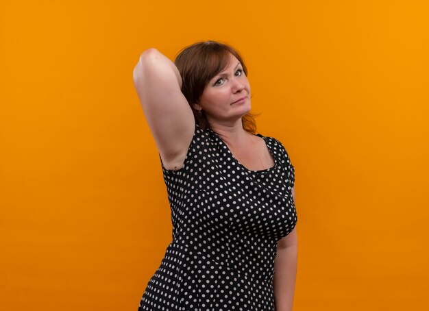 Seriously looking middle-aged woman putting hand behind on isolated orange wall with copy space