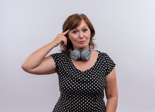 Seriously looking middle-aged woman putting her finger on temple with headphones around her neck on isolated white wall