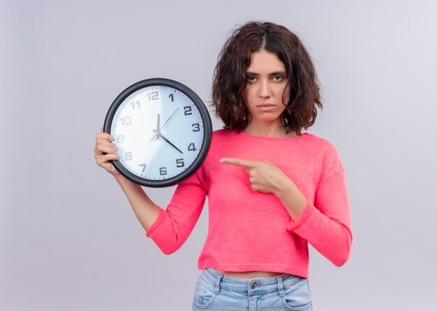 Seriously looking young beautiful woman holding clock and pointing at it on isolated white wall