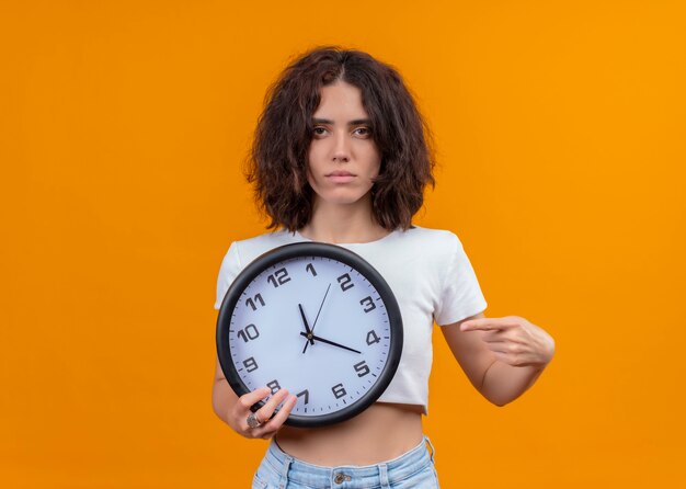 Seriously looking young beautiful woman holding clock and pointing with finger at it on isolated orange wall with copy space