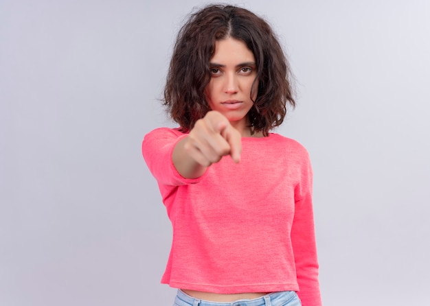 Seriously looking young beautiful woman pointing on isolated white wall with copy space