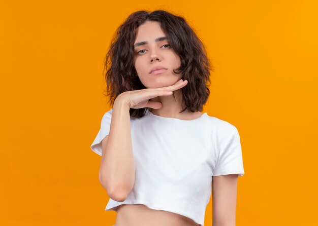 Seriously looking young beautiful woman putting hand under her chin on isolated orange wall with copy space