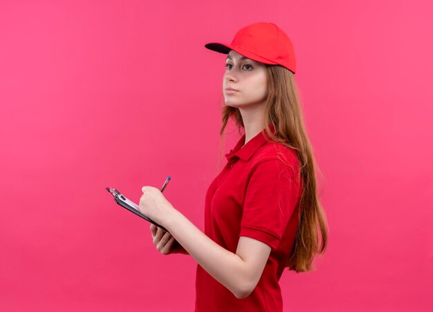 Seriously looking young delivery girl writing on clipboard looking at left side standing in profile view on isolated pink space with copy space