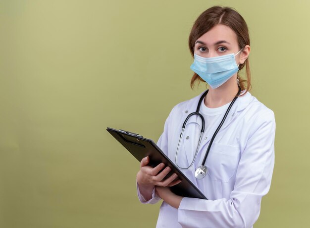 Seriously looking young female doctor wearing medical robe, mask and stethoscope holding clipboard on isolated green space with copy space