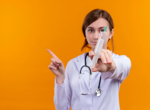 Seriously looking young female doctor wearing medical robe and stethoscope holding syringe and pointing at left side on isolated orange space with copy space