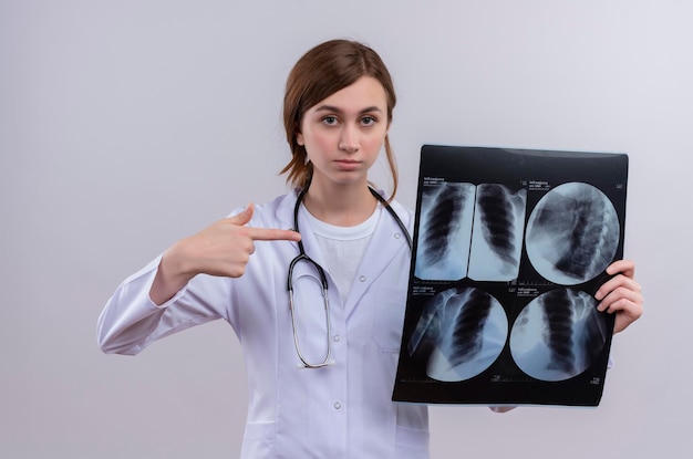 Seriously looking young female doctor wearing medical robe and stethoscope and holding x-ray shot pointing at it 