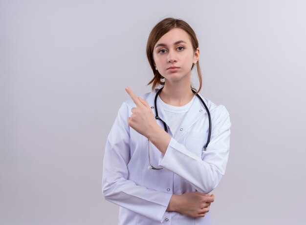 Seriously looking young female doctor wearing medical robe and stethoscope pointing at left side  with copy space