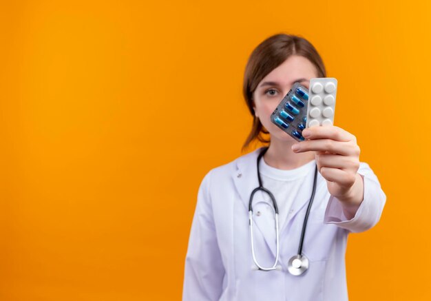 Серьезно выглядящая молодая женщина-врач в медицинском халате и стетоскопе растягивает медицинские препараты на изолированном оранжевом пространстве с копией пространства