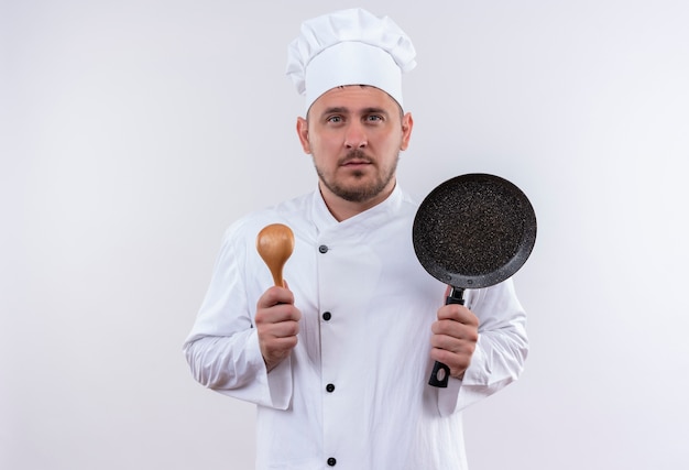 Seriously looking young handsome cook in chef uniform holding spoon and frying pan looking  on isolated white space