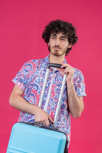 Seriously looking young handsome curly traveler man holding suitcase on isolated pink space