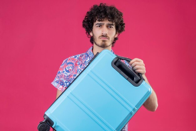 Seriously looking young handsome curly traveler man holding suitcase looking  on isolated pink space