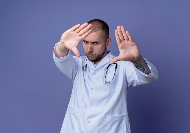 Seriously looking young male doctor wearing medical robe and stethoscope around his neck stretching out hands towards front isolated on purple wall