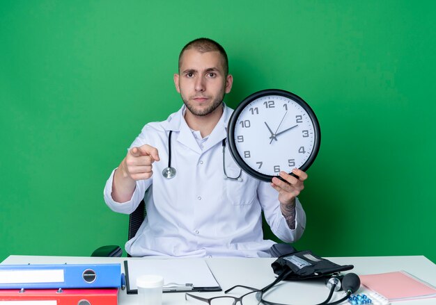 Seriously looking young male doctor wearing medical robe and stethoscope sitting at desk with work tools holding clock and pointing at front isolated on green wall