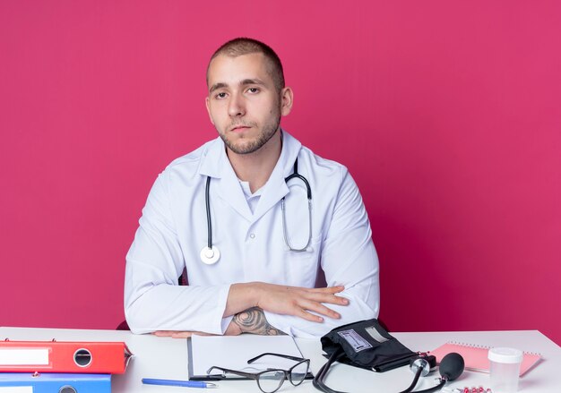 ピンクの壁に隔離された正面を見て机の上に手を置く作業ツールと机に座って医療ローブと聴診器を身に着けている真剣に見える若い男性医師