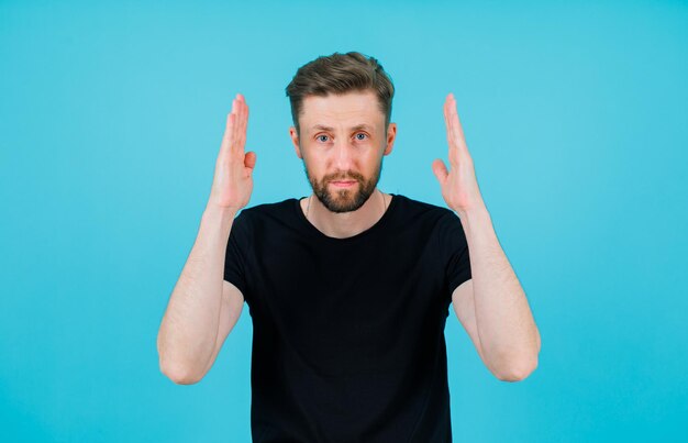 Seriously man is looking at camera by holding hands near head on blue background