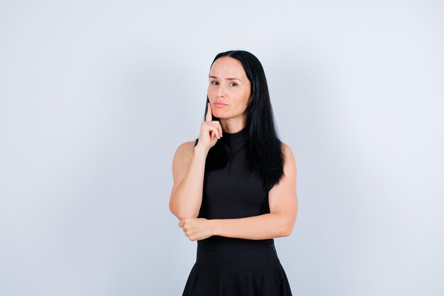 Seriously young girl is looking at camera by holding forefinger on cheek on white background