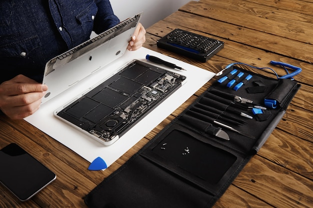 サービスマンは、コンピュータのラップトップの裏側のトップケースカバーを開いてから、木製のテーブルの近くにあるツールキットボックスからプロのツールを使用して修理、クリーニング、修理します。