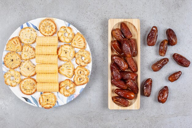 A serving of biscuits on a painted platter with a pile of dates on and next to a wooden tray on marble surface