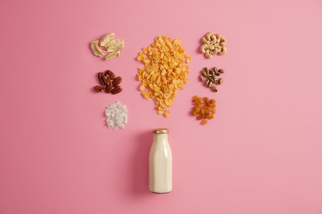 Set of cereals, pistachio, raisins, pecan nuts, dried apple, cashew, coconut around bottle of milk isolated on pink background. Nourishing breakfast rich in vitamins to consume, nutrition concept.