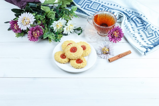 Set of cinnamon, cup of tea, kitchen towel and cookies, flowers on a white wooden board background. high angle view.