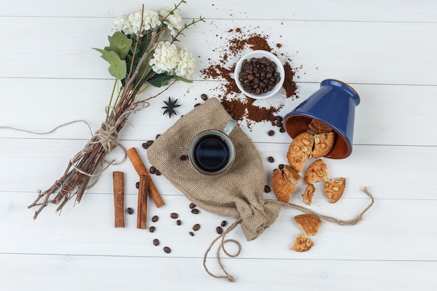 Набор кофейных зерен, печенья, цветов, палочек корицы и кофе в чашке на фоне деревянных и мешков. вид сверху.
