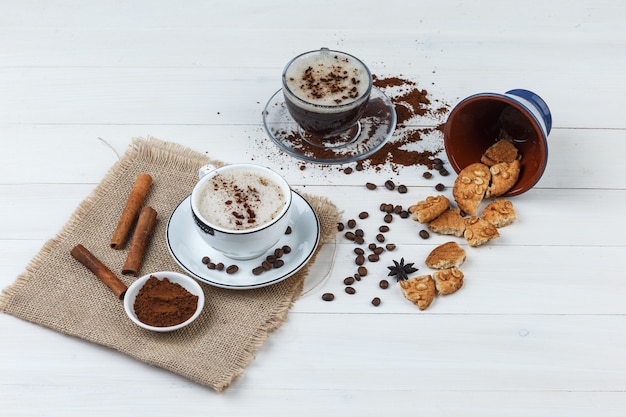 Set of coffee beans, grinded coffee, cookies, cinnamon sticks and coffee in cups on wooden and piece of sack background. high angle view.