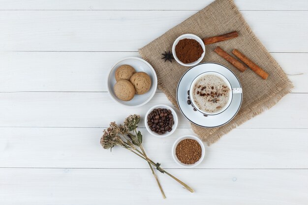 Набор кофейных зерен, молотого кофе, специй, печенья, сушеных трав и кофе в чашке на деревянном фоне и кусок мешка. вид сверху.