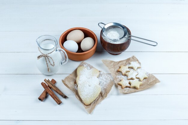 Set of cookies, cinnamon sticks, milk, sugar powder and eggs in a bowl on a wooden background. high angle view.
