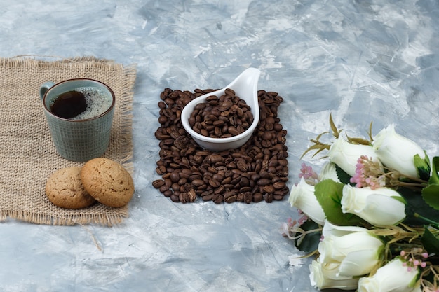 Set of cookies, cup of coffee, flowers and coffee beans in a white porcelain jug on a blue marble background. close-up.
