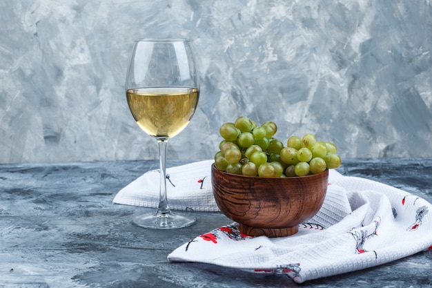 Free photo set of a glass of wine and green grapes in a bowl on grungy plaster and kitchen towel background. side view.