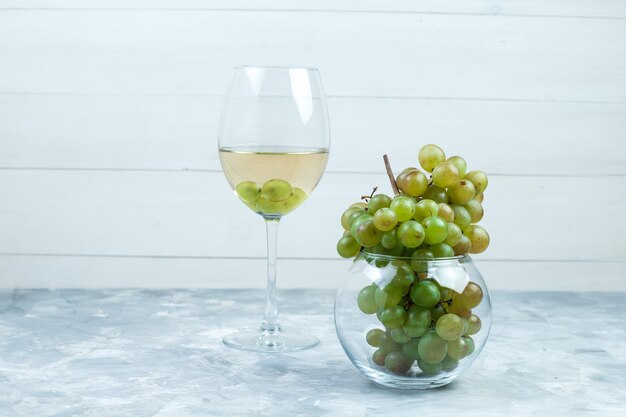 汚れた灰色と木製の背景のガラスの鍋にワインと緑のブドウのガラスのセットです。側面図。