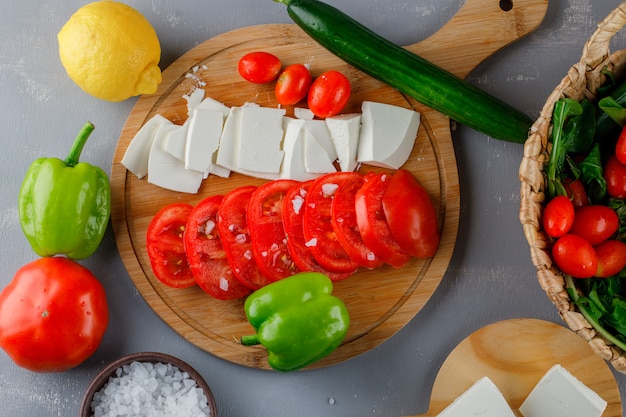 Set of green pepper, lemon, cucumber, salt and sliced cheese and tomatoes on a cutting board on a gray surface