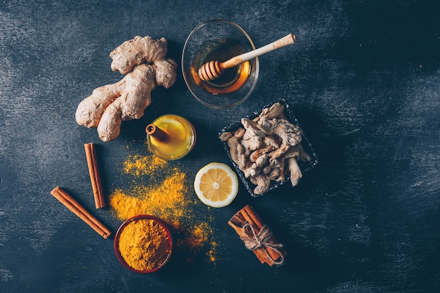 Set of honey, lemon, ginger and dry cinnamon pack and ginger powder in bowls on a dark textured background. top view.