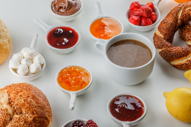 Set of jams, raspberry, sugar, chocolate in cups, turkish bagel, bread, lemon and a cup of coffee on a white surface
