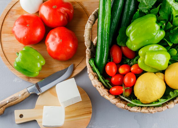 set of knife, green pepper, lemon, cucumber, cheese, greens and tomatoes in a cutting board on a gray surface