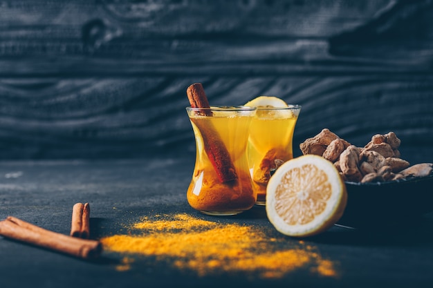 Free photo set of lemon, ginger and dry cinnamon and ginger powder in a tea cups on a dark textured background. side view. space for text