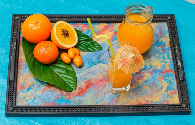 Бесплатное фото Набор сока в очках, листья, апельсин мандарина и апельсины в рамке с абстрактными цветами на голубой. высокий угол обзора.