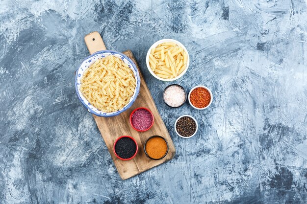 Set of spices and assorted pasta in bowls on grey plaster and cutting board background. top view.