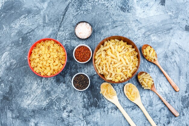 Free photo set of spices and assorted pasta in bowls and wooden spoons on a grungy plaster background. flat lay.