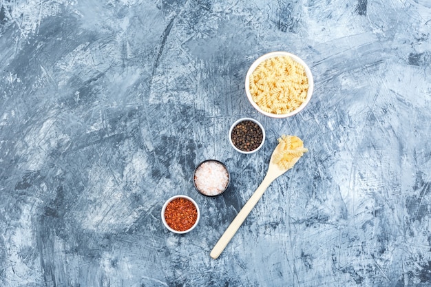 Set of spices and fusilli pasta in white bowl and wooden spoon on a grungy plaster background. flat lay.