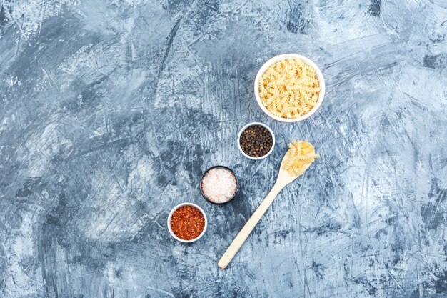 汚れた石膏の背景に白いボウルと木のスプーンでスパイスとフジッリパスタのセット。フラットレイ。