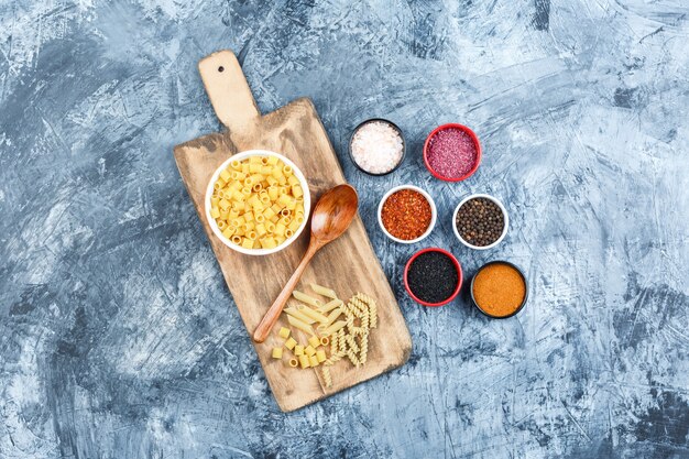 Set of spices, wooden spoon and ditalini pasta in a bowl on grey plaster and cutting board background. top view.