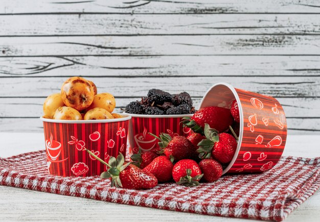 Set of strawberries, loquats and mulberries in a bowls on a cloth and light wooden background. side view.