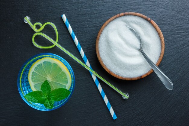 Set of straws, leaves and spoon with salt and lemon slice in a blue bowl on a dark stone surface. top view.