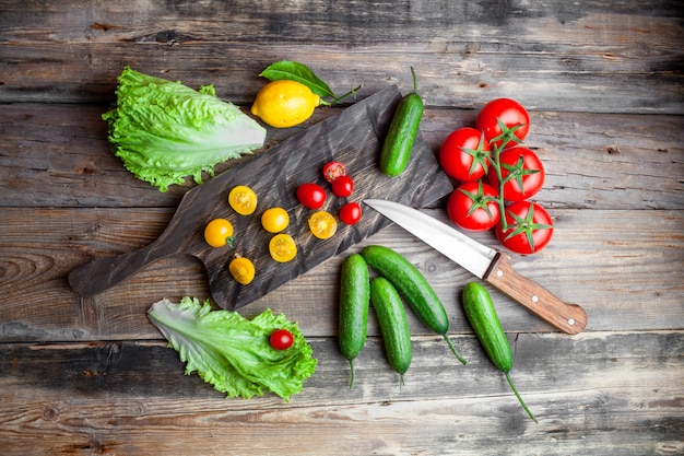 Set of tomatoes, lettuce, lemon and knife and cucumber in a cutting board on a dark wooden background. top view.