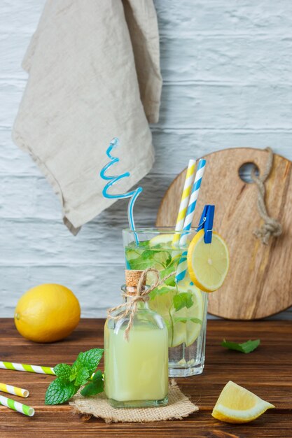 Set of wooden crate and lemons and white cloth, cutting board and glass of lemon juice on a wooden and white surface. side view.