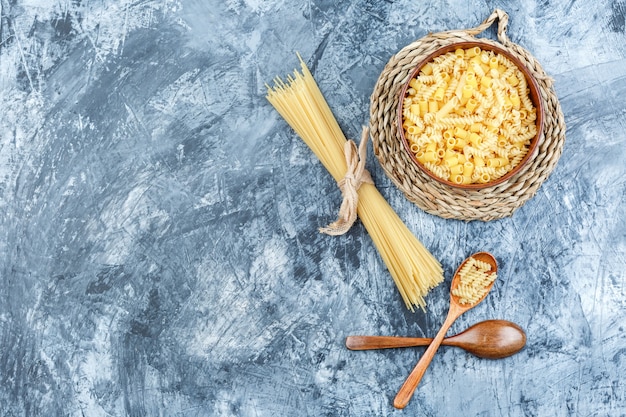 Set of wooden spoons and assorted pasta in a bowl on grey plaster and wicker placemat background. top view.