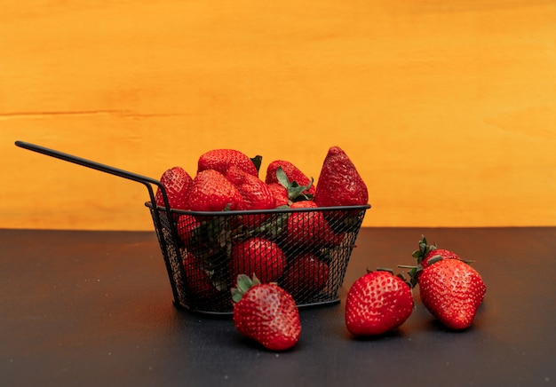 Several strawberries in a black basket on a orange background. side view.